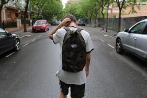 Teenager walking in the middle of a road away from camera, wearing a back back, scratching the back of their head
