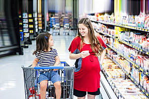 A pregnant woman in a supermarket aisle. Her young daughter is next to her sat in a supermarket trolley