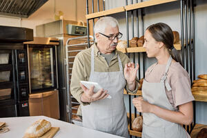 Mature French baker holding tablet giving instructions to younger female colleague