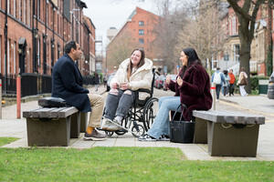 Visually impaired man chatting with a lady in a wheelchair and another lady in an open space.