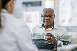 An elderly man looks at a pill bottle in his hand while sitting across the table from a doctor in the doctor's office.