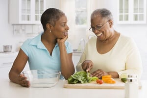 A young woman on the left holds a glass bowl while speaking to an older woman on her right chopping vegetables on a chopping board.