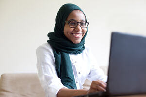 A woman in a dark blue hijab and white shirt sits on her laptop smiling. 