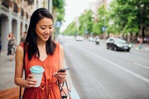 A woman with dark hair and an orange dress holds her smartphone and a cup of coffee next to a road full of cars.