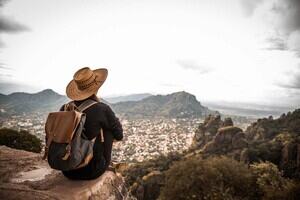A woman sat on a rocky mountain wearing a large rimmed hat and rucksack, starring out over a town below. 