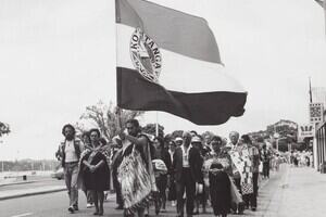 A black and white photograph of Māori men and women. one man and women are in traditional Māori dress, the man is holding a flag. They are in a crowd and are looking down, in prayer.