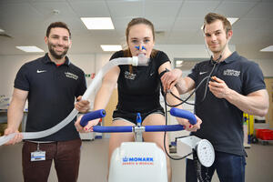Woman on a stationary bike performing a fitness test with two male sport and exercise scientists from The University of Hull either side.