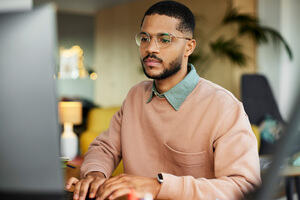 A man in a pink jumper and glasses is on a computer in an office setting.