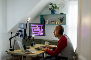 Young man wearing a headset sitting at his desk and playing a game on one of two computer screens.