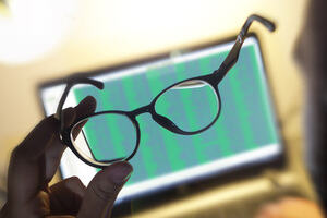 A man holds his glasses in front of a computer screen with a spreadsheet on it.
