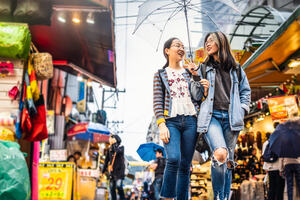 Two happy asian girls holding an umbrella and walking in a street market.